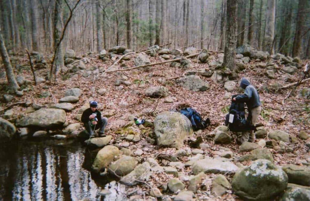 Two hikers sitting near stream and filtering water before their hike.